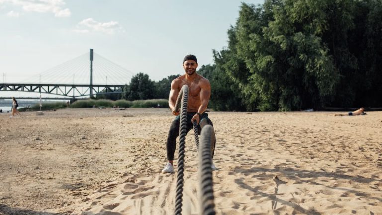 A muscular adult male with battle ropes exercises on a sunny beach by a bridge.